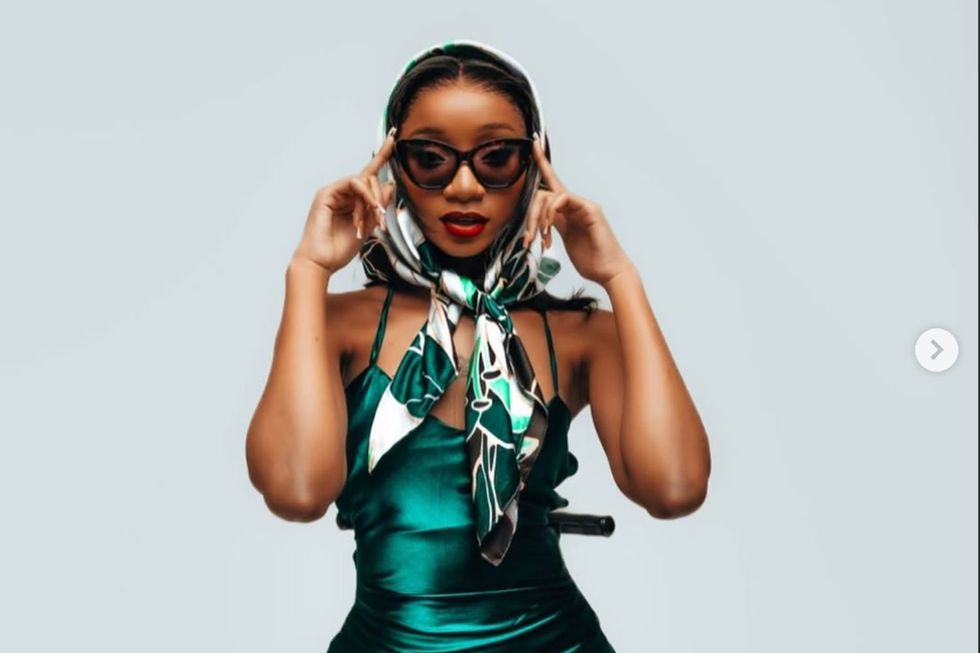 Ajib Gathoni poses with shades. She wears a teal green dress and a babushka on her head.