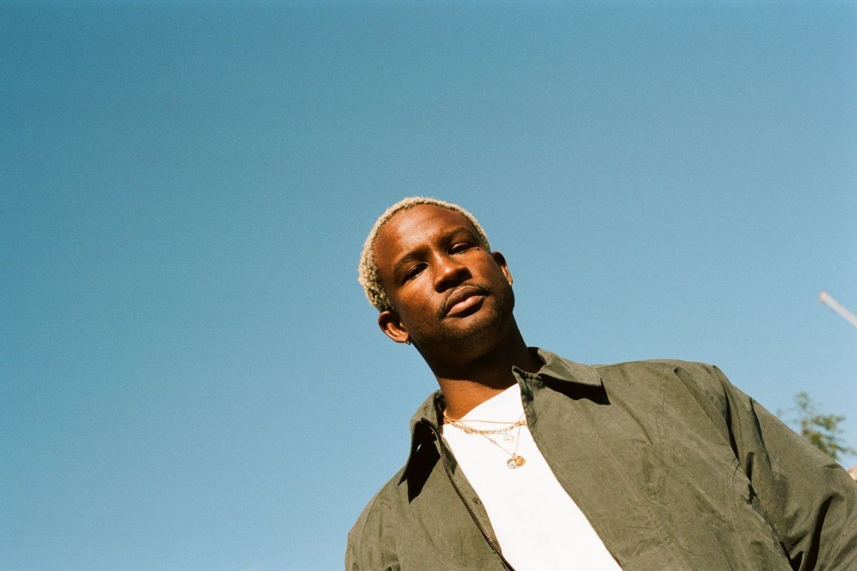 Akinola Davies Jr. wearing a white shirt under a green jacket, with short blond hair and layered gold necklaces, standing against a clear blue sky.