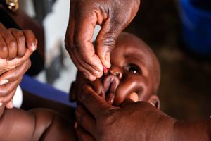 An adult hand is seen giving a baby medication in its mouth, while another adult hand holds the baby's hand.