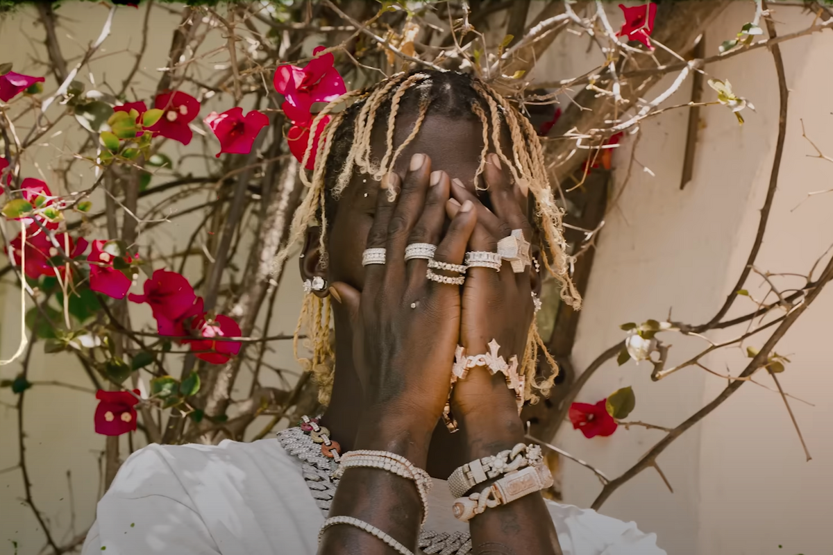 An artist holds his hand in front of his face, revealing extensive jewelry on his hands and neck, in a new music video for Nigerian artist Olamide.
