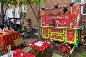 An Egyptian food cart, painted in red, white, and green, in the front yard of a building, surrounded by red tables and chairs. 