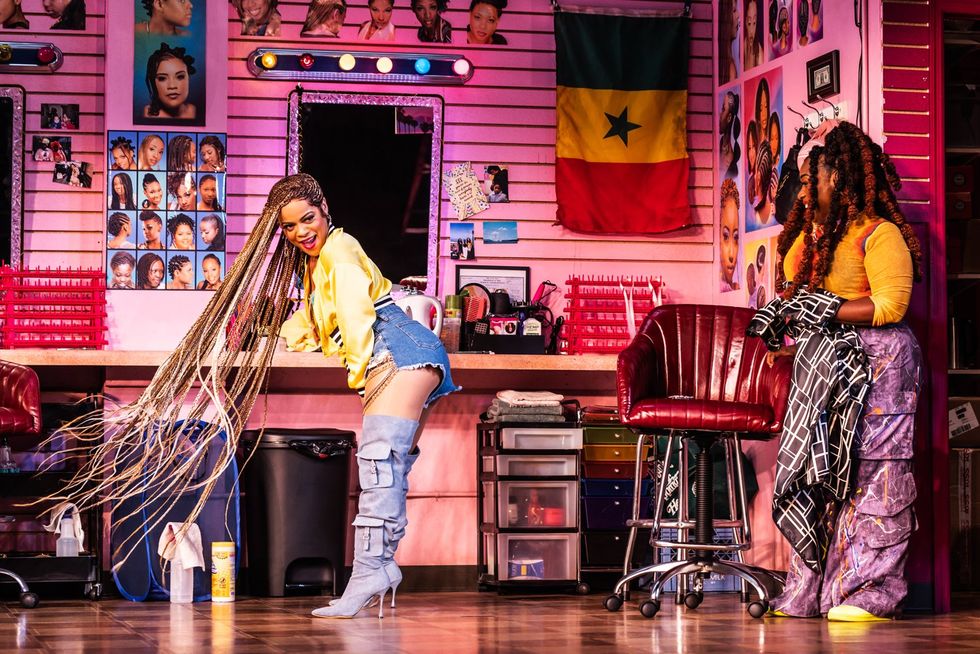 An image from the play of two women in a hair braiding shop, one is showing off her long braids while the other looks on in glee.