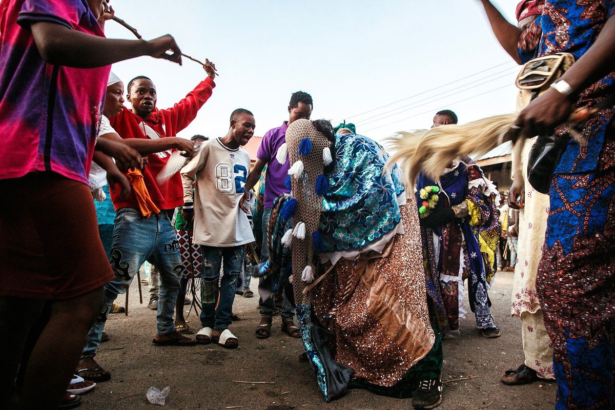 An image of a group of men at Nigeria's Egungun Festival, with sticks circling another man who is wearing colorful regalia and dressed up as an ancestor. 