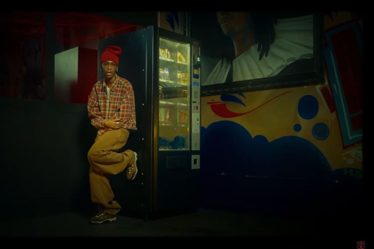 An image of K.Keed from the “Legwork” video. She is resting one leg on a vending machine, wearing a red beanie and a checked shirt.