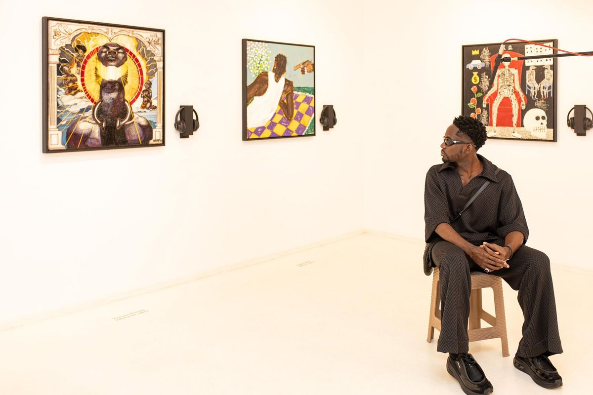 An image of Mr Eazi sitting on a chair in a gallery, looking back at three artworks hanging on the wall. 