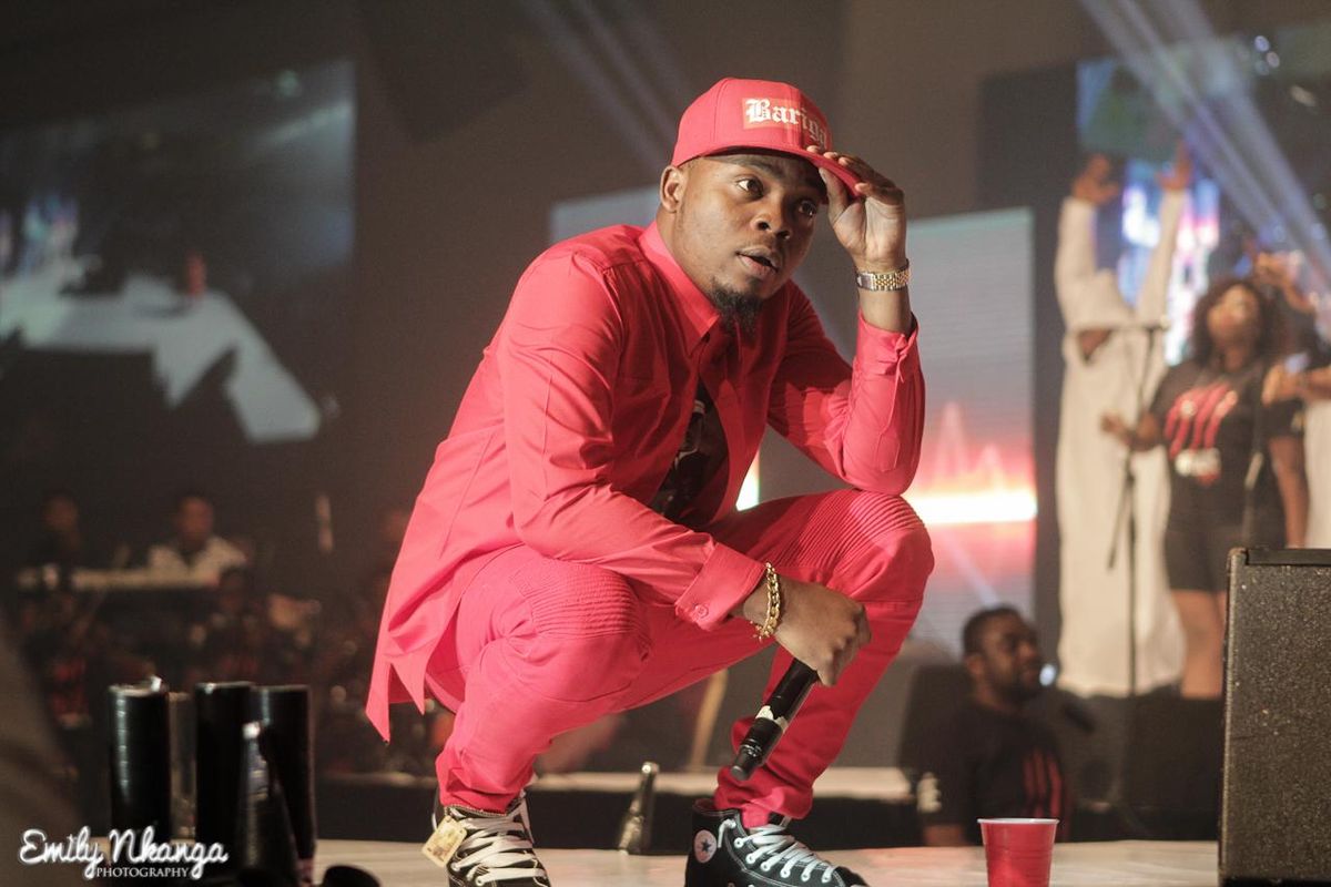 An image of Olamide dressed in red, crouching on a stage, holding a microphone in his hand