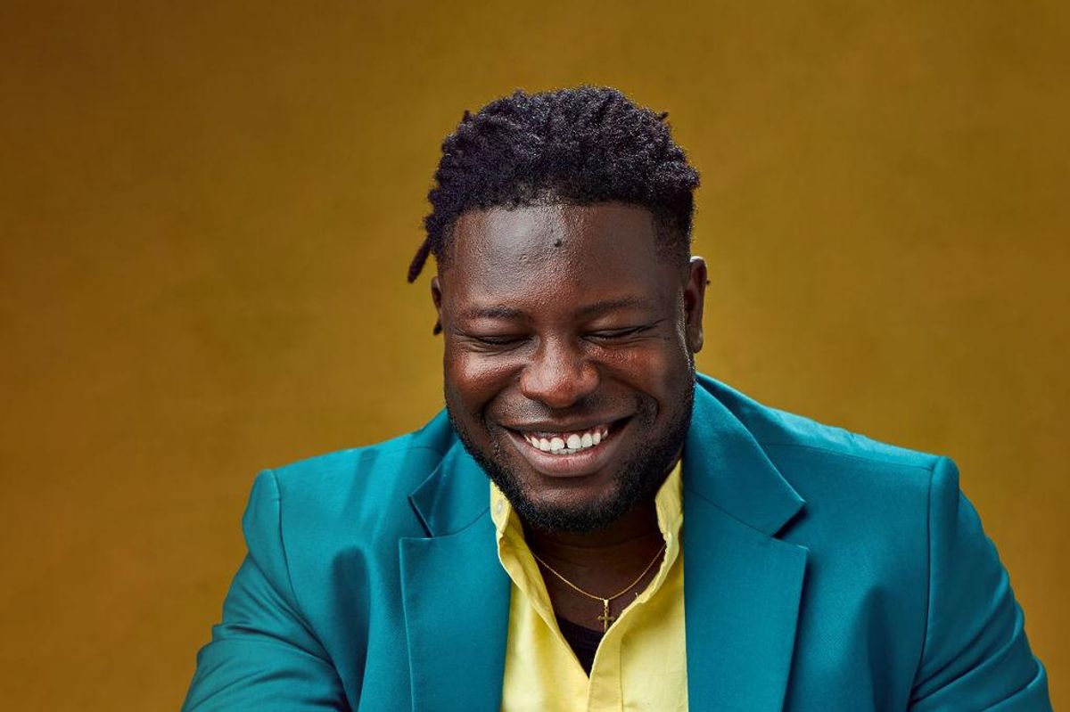 An image of Temitope Dada smiling against a mustard-colored background, wearing a blue suit.