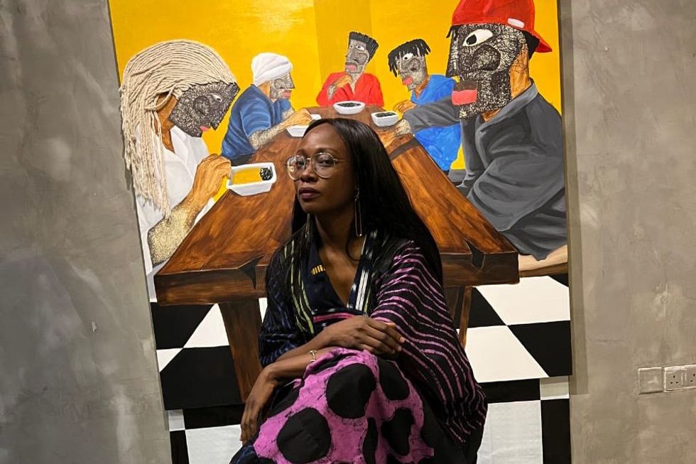 An image of the designer sitting on a chair that\u2019s placed on a checkered floor and there\u2019s a vibrant art piece behind her.