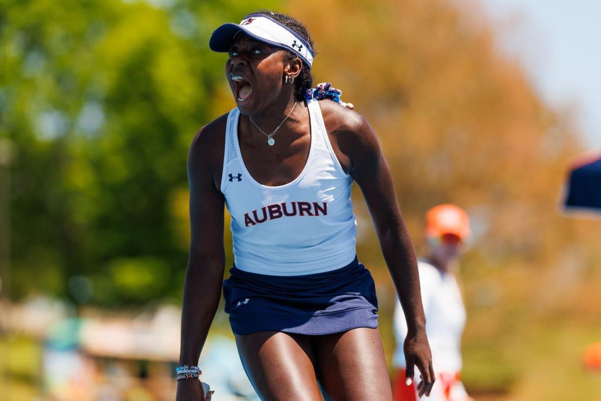 Angella Okutoyi wearing an Auburn University tennis uniform, shouting in celebration during a match, with a blurred outdoor background and a visible official in the distance.
