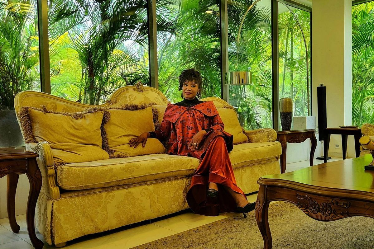 Ashionye Raccah poses while sitting on a couch behind a large glass window overlooking lush greenery.