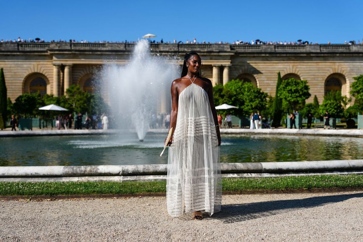 Aya Nakamura wears a sheer off-white halter-neck gown featuring vertical textured stripes and geometric horizontal embroidery across the skirt, styled with metallic open-toe heels, a white folding hand fan, and minimal jewelry.
