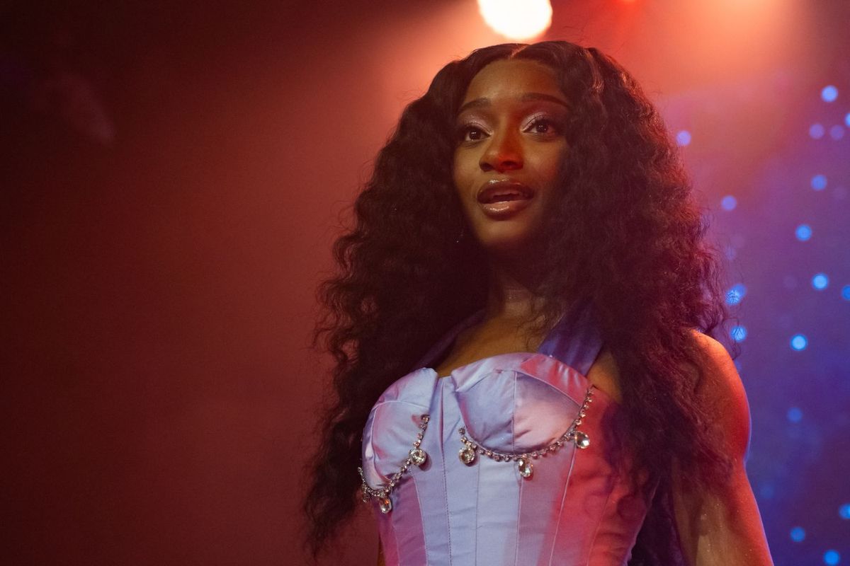 Ayra Starr stands on stage under warm red and purple lighting, wearing a lavender satin corset adorned with rhinestones, her long, voluminous hair cascading in waves over her shoulders as she gazes slightly upward with a poised expression.