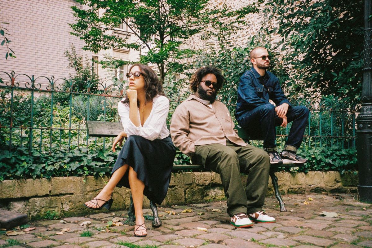 Bas and The Hics are sitting on a wooden bench in a London backyard, all wearing shades and appearing to look in different directions.