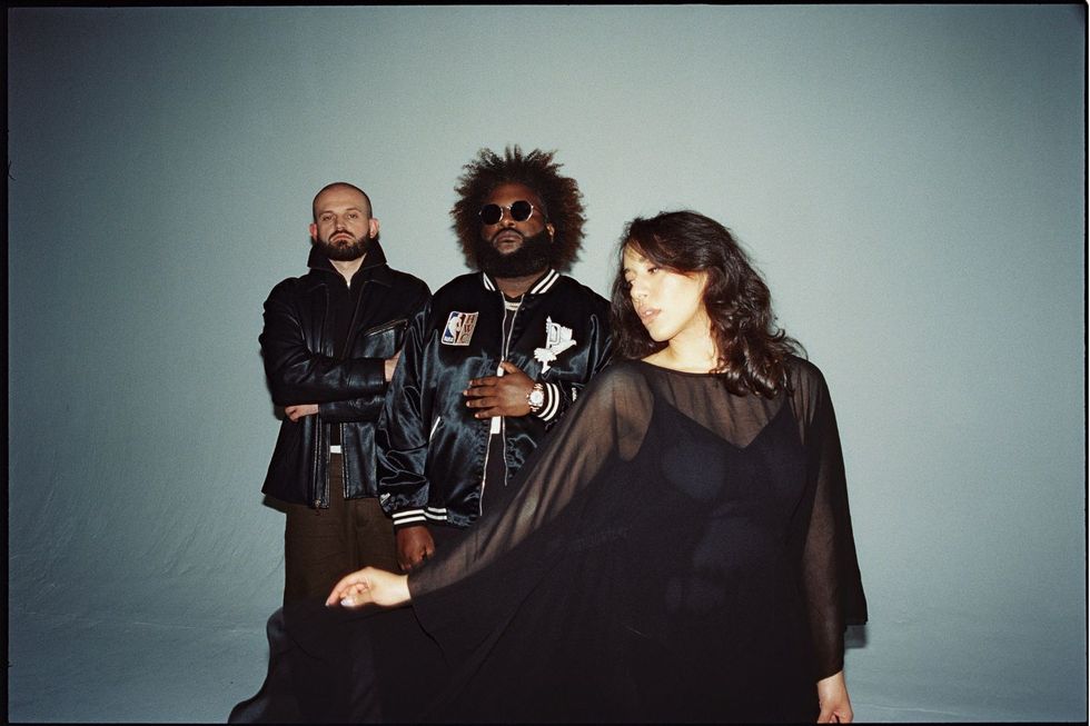 Bas and The Hics in front of a blue-grey background, wearing all black. Barker is standing in front of the two men and holding her dress up to the side while Bas and Evans look into the camera.