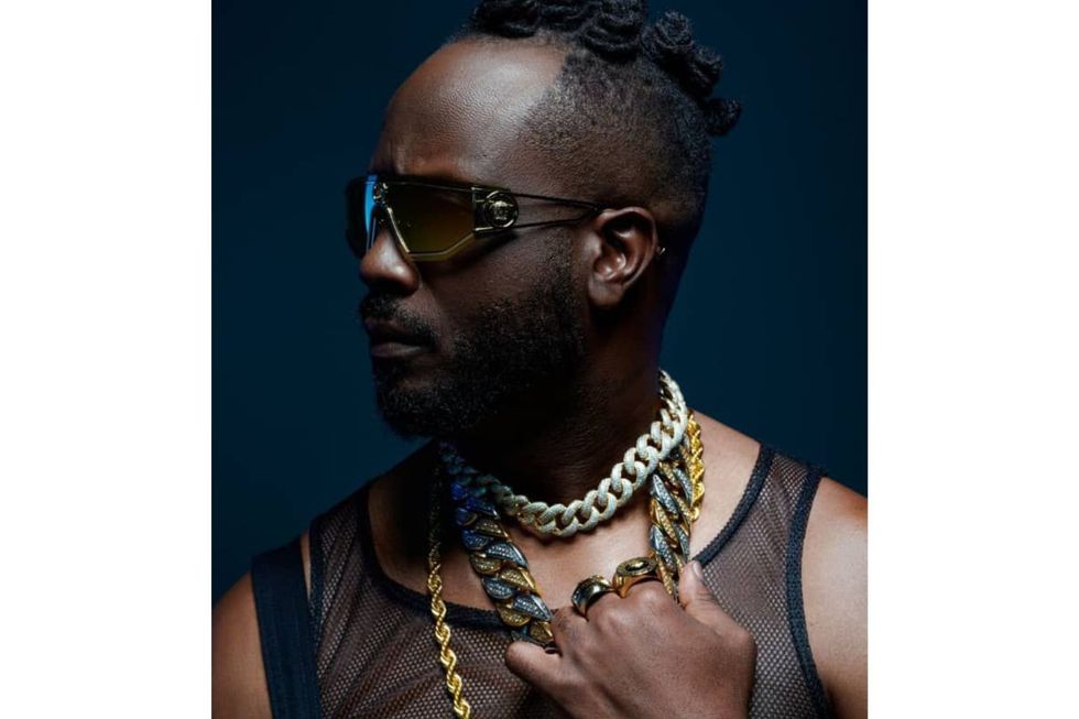 Bebe Cool wearing layered gold chains and futuristic sunglasses, looking sideways against a dark background.
