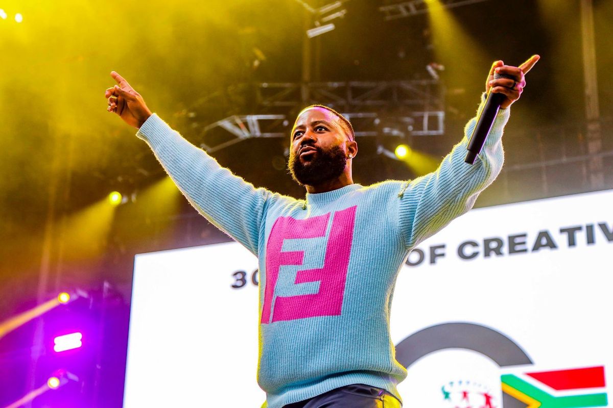 Cassper Nyovest on a stage, mic in one hand, hands stretched out, wearing a blue top with a pink Fendi logo on it.