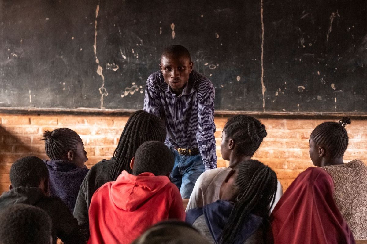 Charles Lipanda stands at the front of a classroom, speaking to a group of young students seated before him at Dzaleka Refugee Camp.