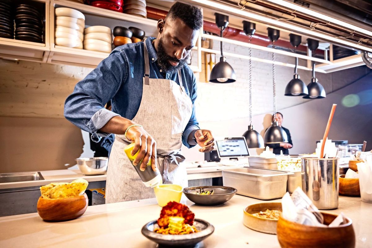Chef Eric Adjepong in a blue shirt and white apron in the kitchen of his new restaurant, Elmina
