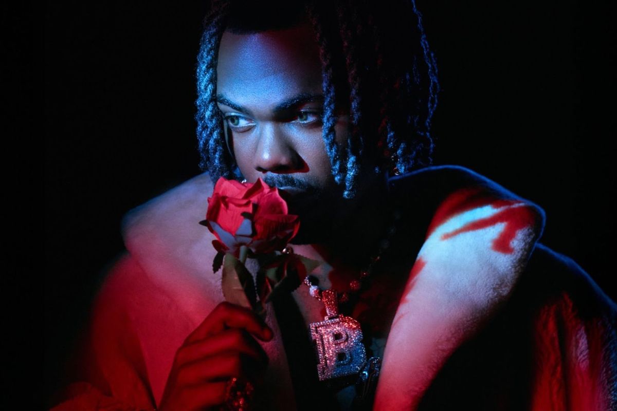 Ckay poses in a dim background for a studio photo while holding a rose close to his nose.