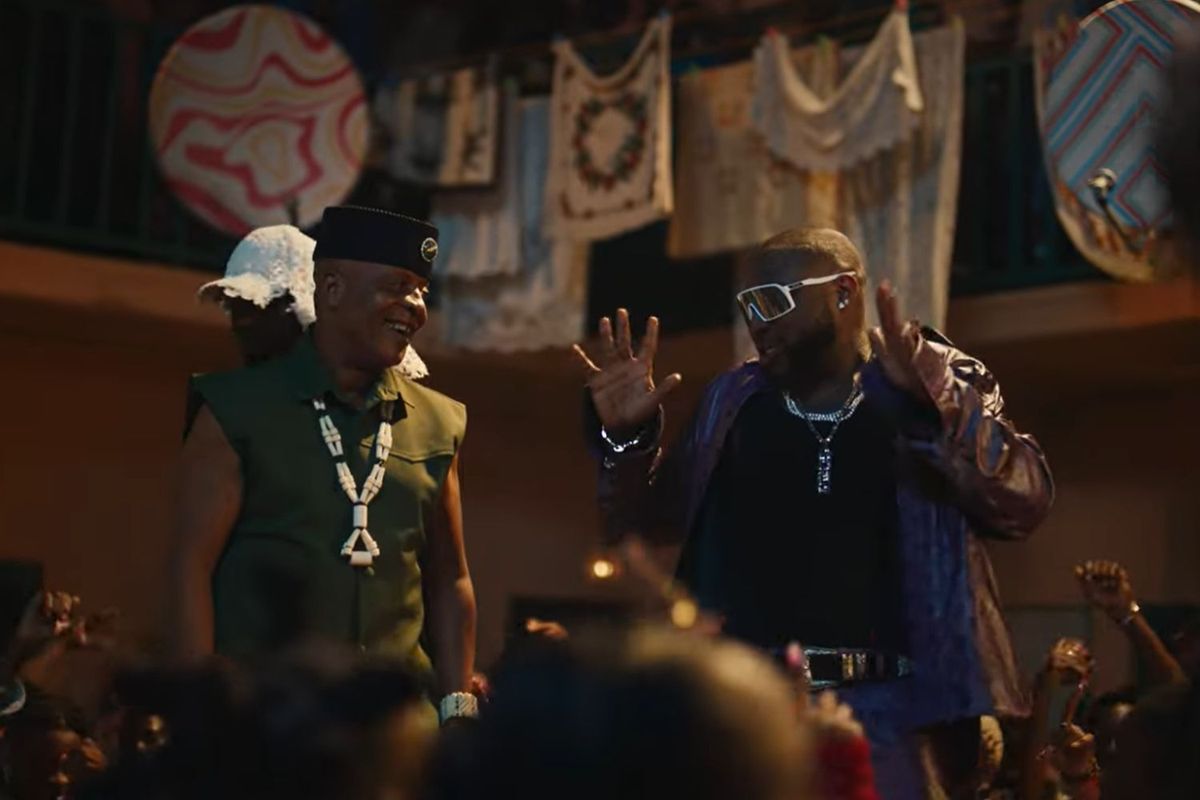 Davido wears a fashionable black jacket and white rimmed sunglasses in the visuals of ‘With You,' he has his hands in the air as he sings and there is a crowd around him.