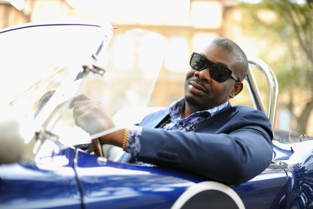 Don Jazzy leans out the side of a blue convertable car wearing a slick blue suit.