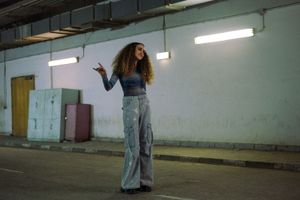 Egyptian singer Donia Wael is standing in a garage, wearing grey trousers, black boots, and a tight shirt with space print. Her long, curly hair is flowing around her smiling face, her right arm is lifted in motion. 