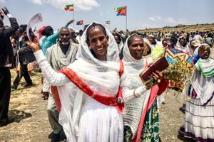 Eritrean women dressed in traditional white habesha clothing celebrate joyfully, waving and holding flowers, as crowds gather behind them. Ethiopian and Eritrean flags fly in the background, symbolizing unity and peace.