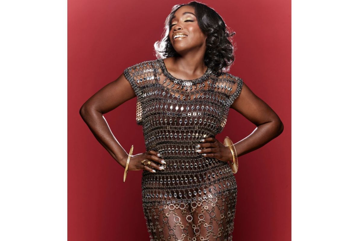 Estelle poses in a chain link dress in front of a red backdrop.