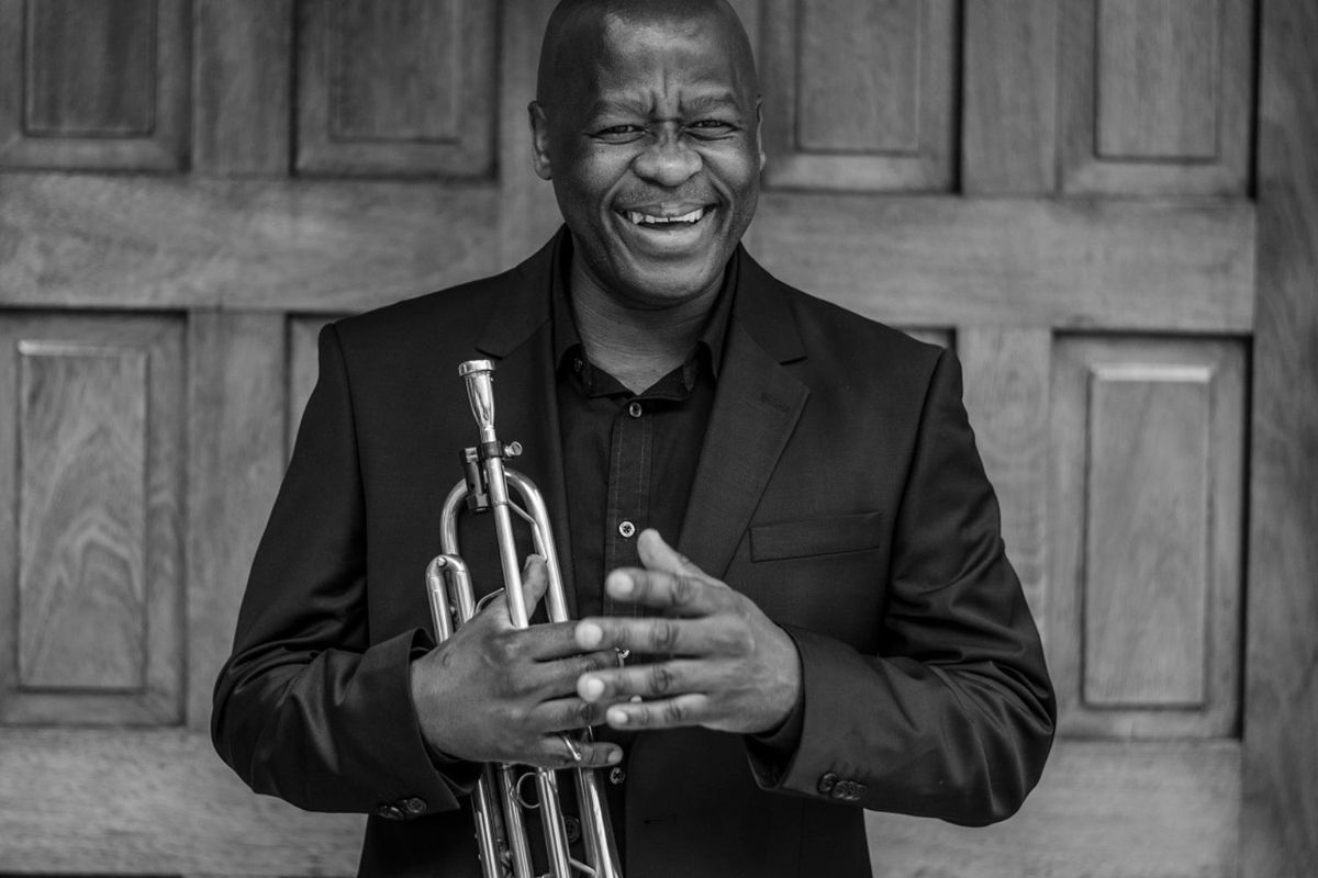 Feya Faku laughs as he poses with his trumpet, wearing a black jacket and a black shirt.