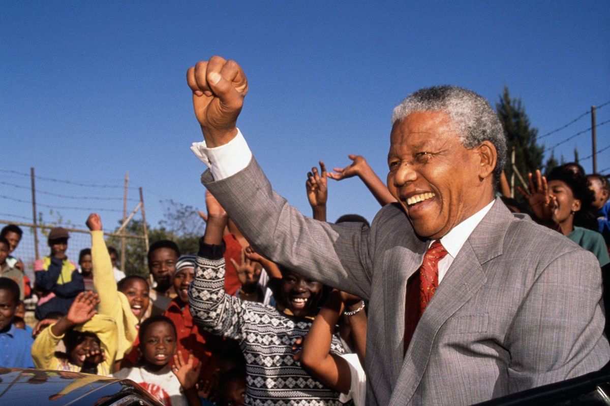 Former South African president Nelson Mandela raises his fist in the air while on a school visit to Hlengiwe School. He spots a grey suit and a white shirt.