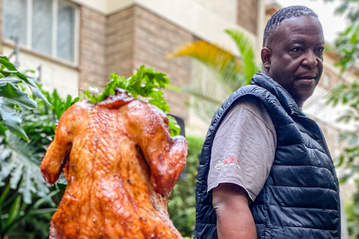 George Kariithi stands next to a roasted beer-can chicken on a wooden board.