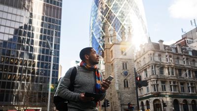 A serious adult black male on his way to a work meeting in London. He is admiring the beautiful streets of London while walking. In his hands, he is carrying a smartphone and a tablet. He looks serious. The weather is nice and sunny. He looks focused.