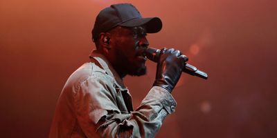 Rapper Stormzy performing with microphone in black hat and beige jacket. 