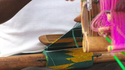 A close up image of kente cloth being woven on a person’s lap.