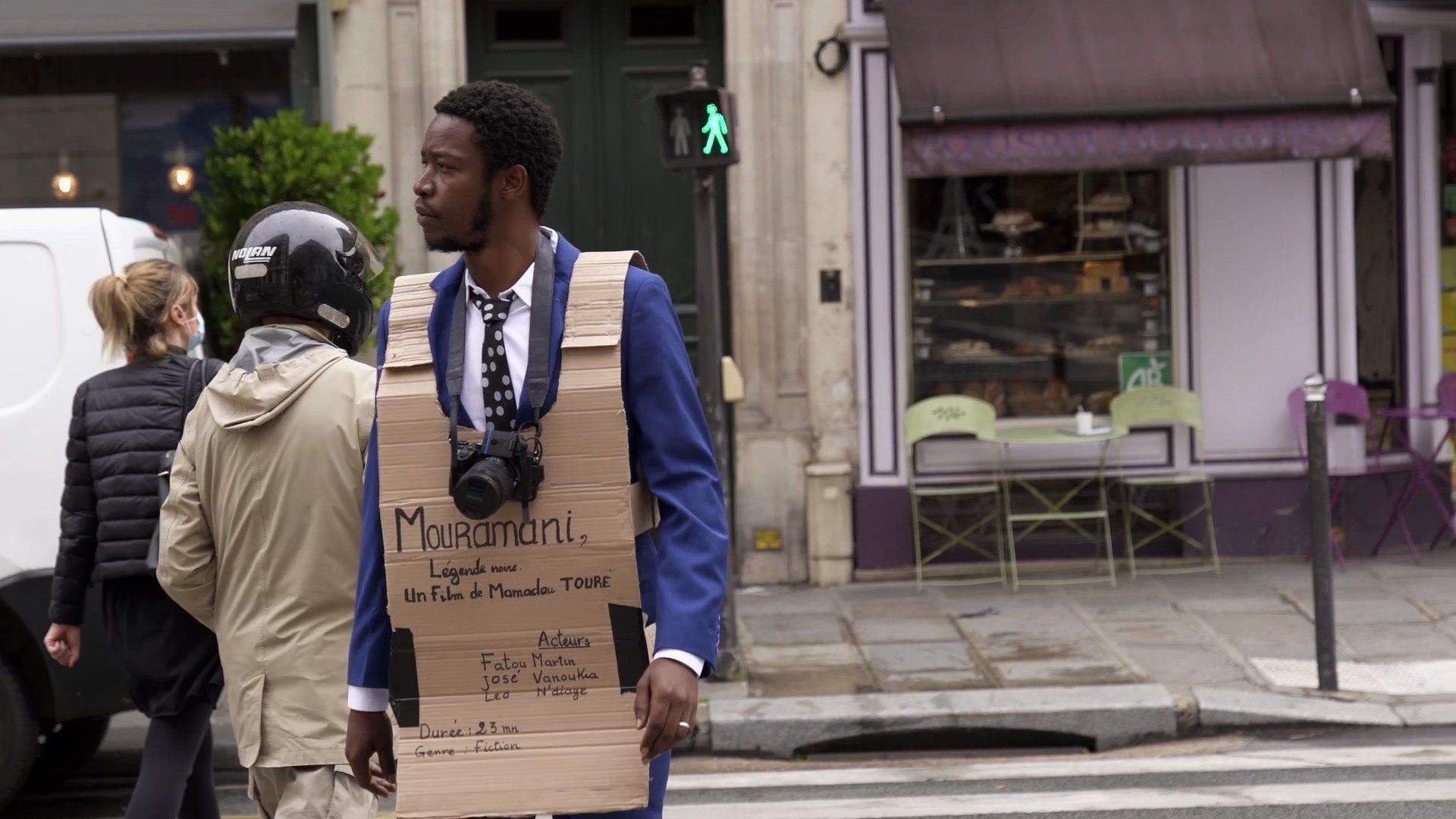 A still image from the film of the director wearing a cardboard cut-out with the film title, Mouramani, written on it and a camera around his neck.