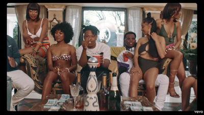 A screenshot from Nasty C's music video for 'Bookoo Bucks' showing him rapping surrounded by scantily dressed women and money. 