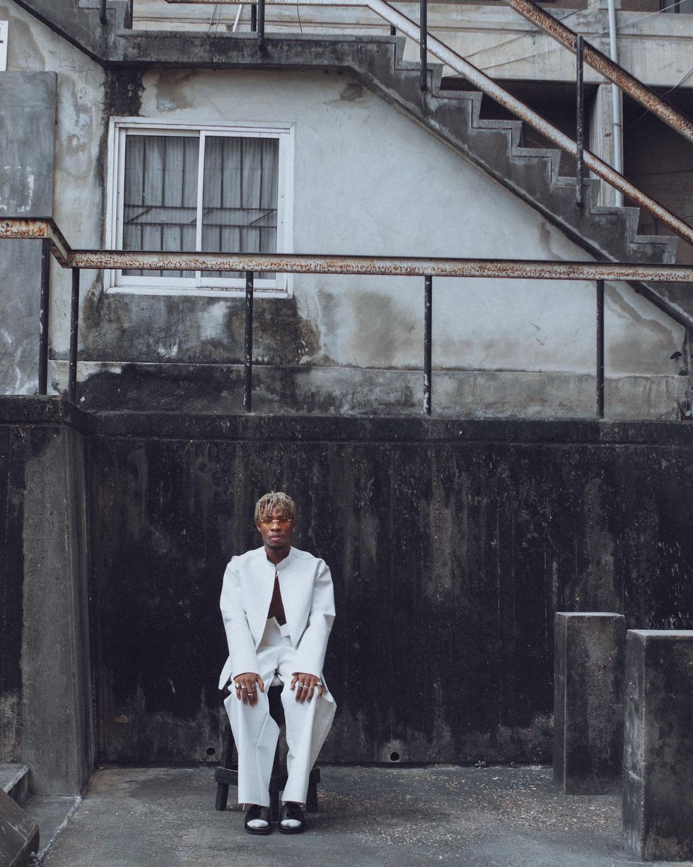 Joeboy sits in a white suit in the street.