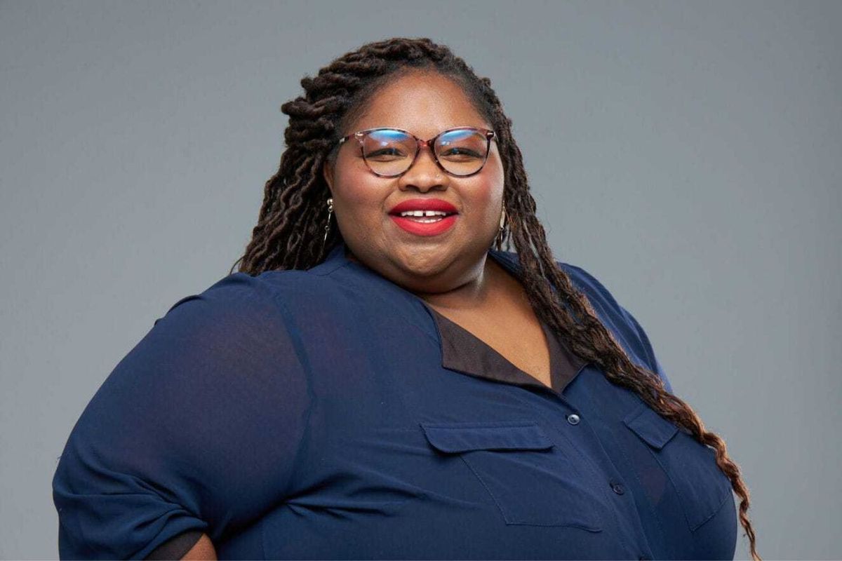 Journalist Tshidi Madia smiles at the camera, wearing a navy top, glasses, and red lipstick