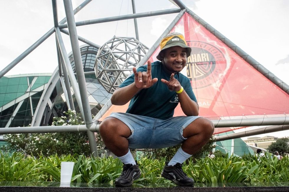 Kabelo Moshims poses for a photo by squating down in jean shorts and a bucket hat.