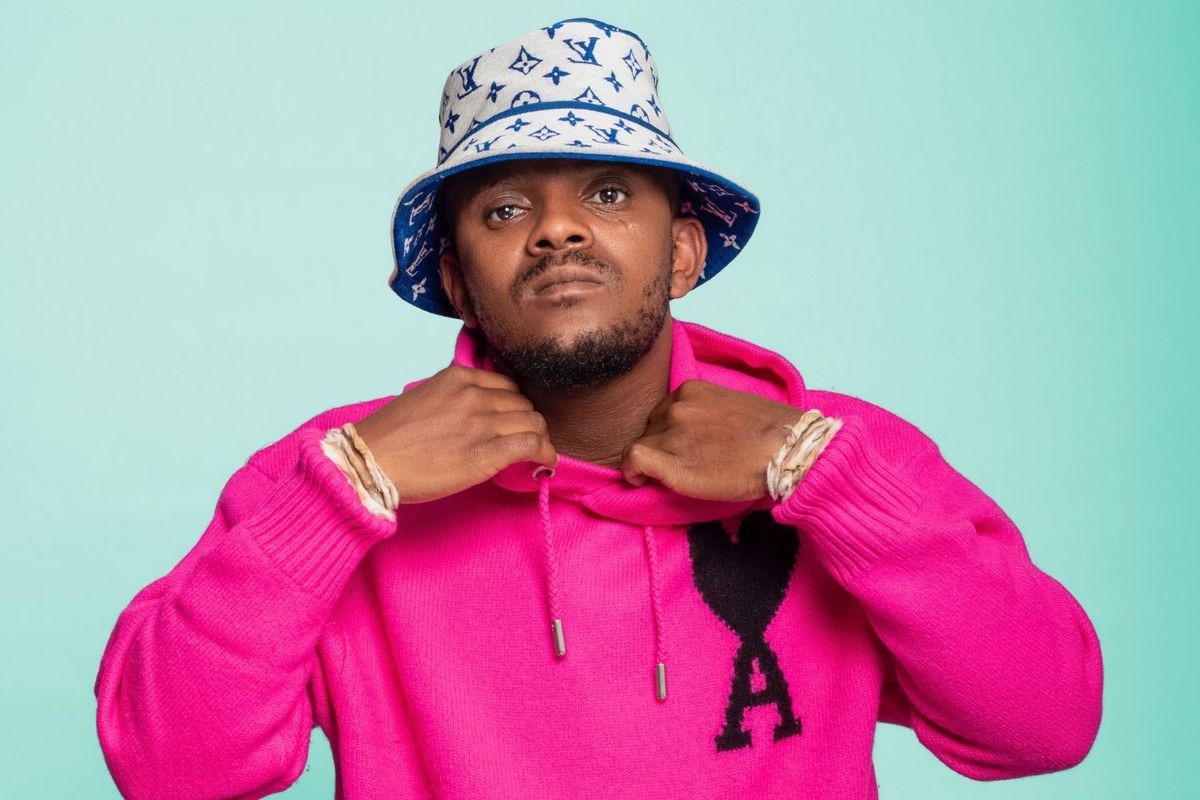 Kabza De Small wears a pink AMI Paris Ami de Coeur wool cardigan. He is also wearing an off-white and blue LV bucket hat, and is holding the collar of his sweater.