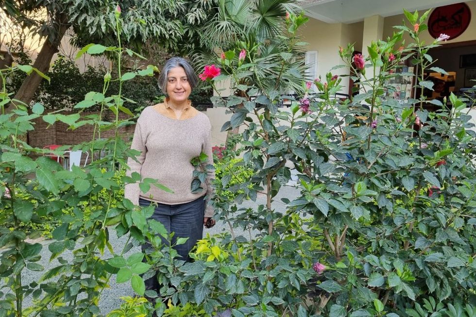 Karam Youssef is standing in front of the entrance to her bookshop, behind a flowerbed with high plants and red flowers. She is wearing a light grey pullover and dark grey trousers.