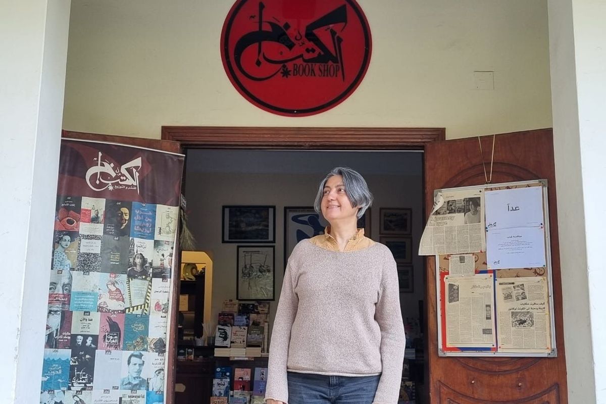 Karam Youssef, wearing a grey pullover, is standing in the entrance of her bookshop. Above her head, a red sign reads “Al Kotob Khan” in Arabic.