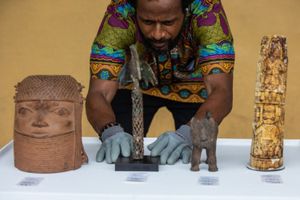 A National Museum exhibition officer in gloves carefully arranges Benin Bronzes on display, including carved ivory, bronze sculptures, and a ceremonial head, highlighting the cultural significance of repatriated African artifacts.
