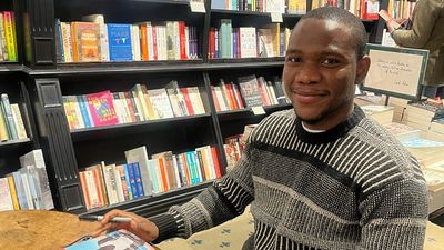 A photo of Chukwuebuka Ibeh sitting in a library with three copies of his book “Blessings” on the table.