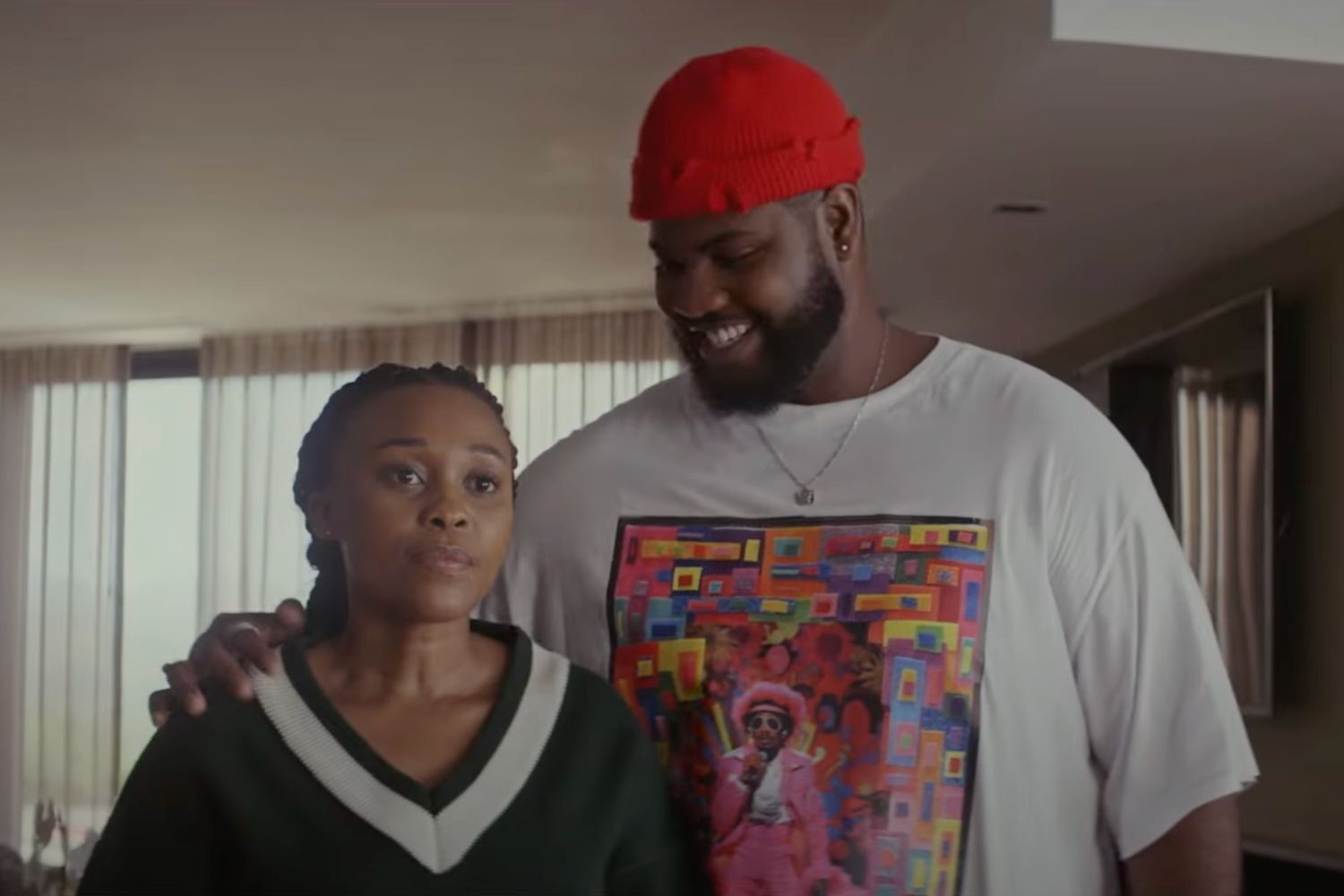 A still from ‘A Scam Called Love’ showing a man in white t-shirt and red beanie resting his hands on and smiling at a woman in a green sweatshirt.