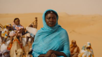 A still image from the film of a woman in the desert in a full headscarf looking pensive. 