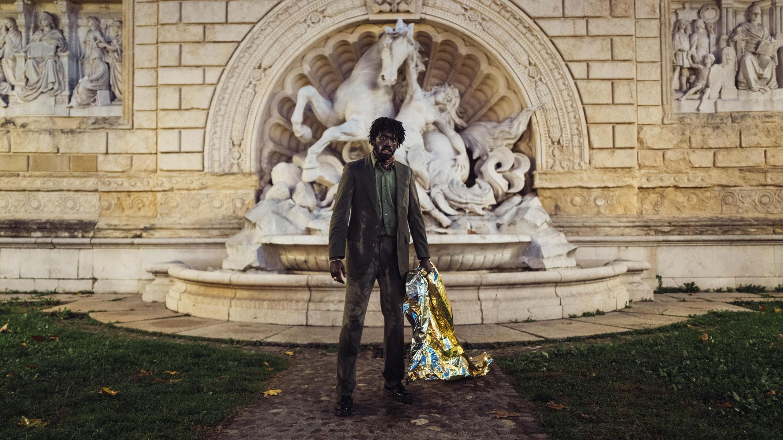 An image from the film, Bravo, Burkina! of a man in a suit standing in front of a fountain with a plastic sheet that he has used to cover himself with. 