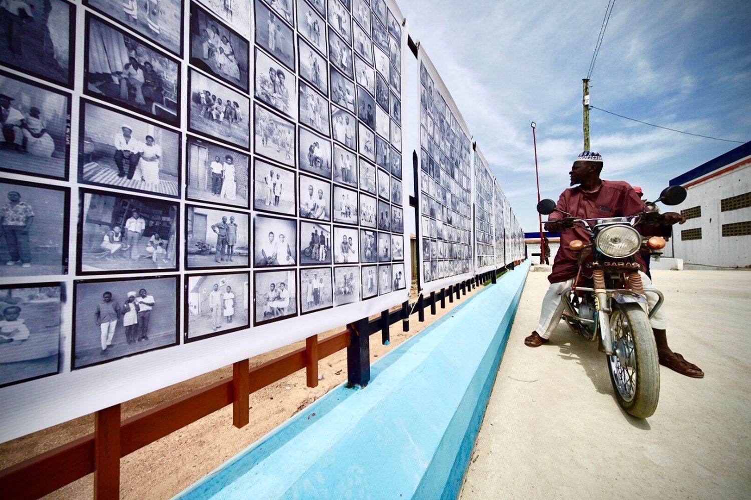 A man on a motorcycle pauses to look at a long outdoor display of black-and-white portrait photographs mounted on a wall under a bright, partly cloudy sky.