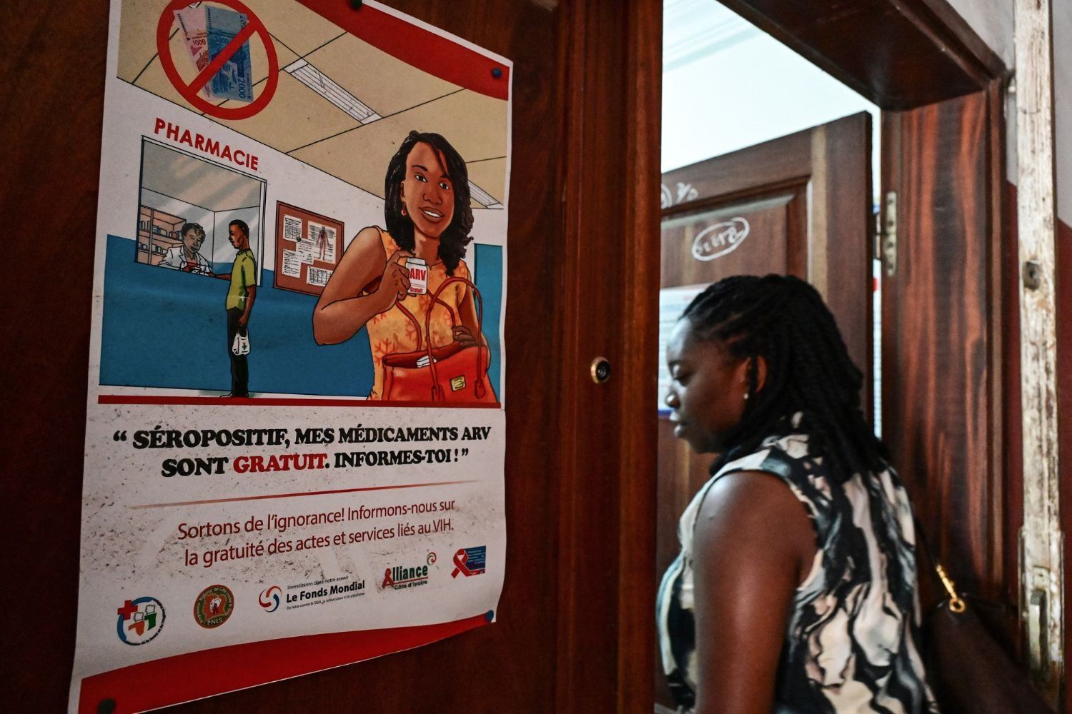 A woman walks past a health awareness poster on a wooden wall, which informs people living with HIV that antiretroviral medications are free, urging them to seek information and support.