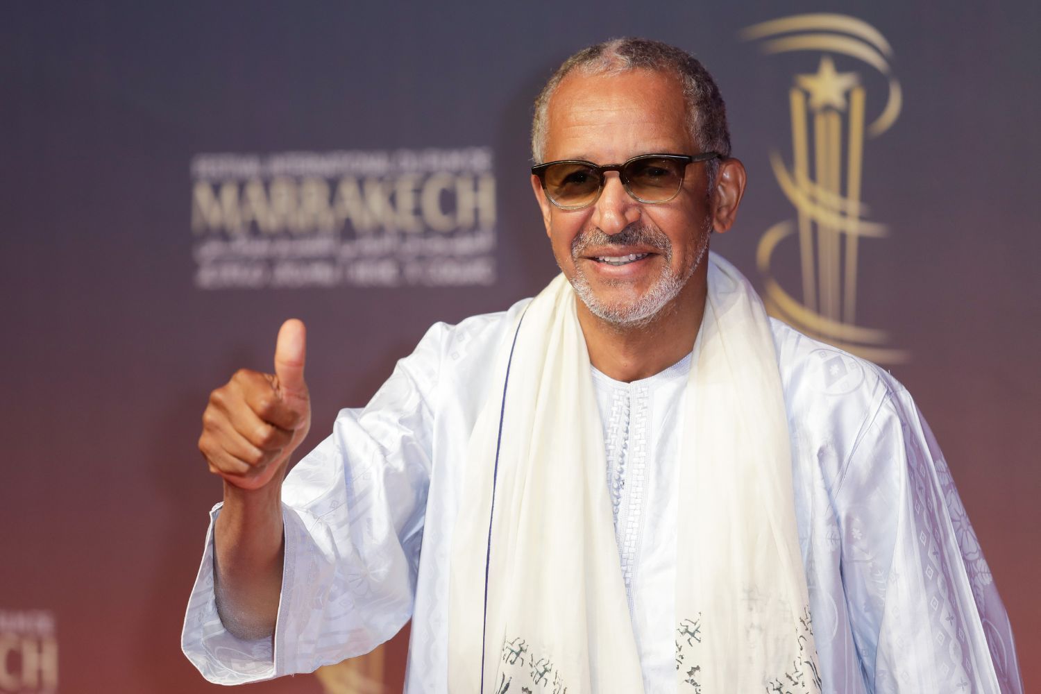 Abderrahmane Sissako dressed in white smiles and gives a thumbs up on the red carpet at the Marrakech International Film Festival in 2024